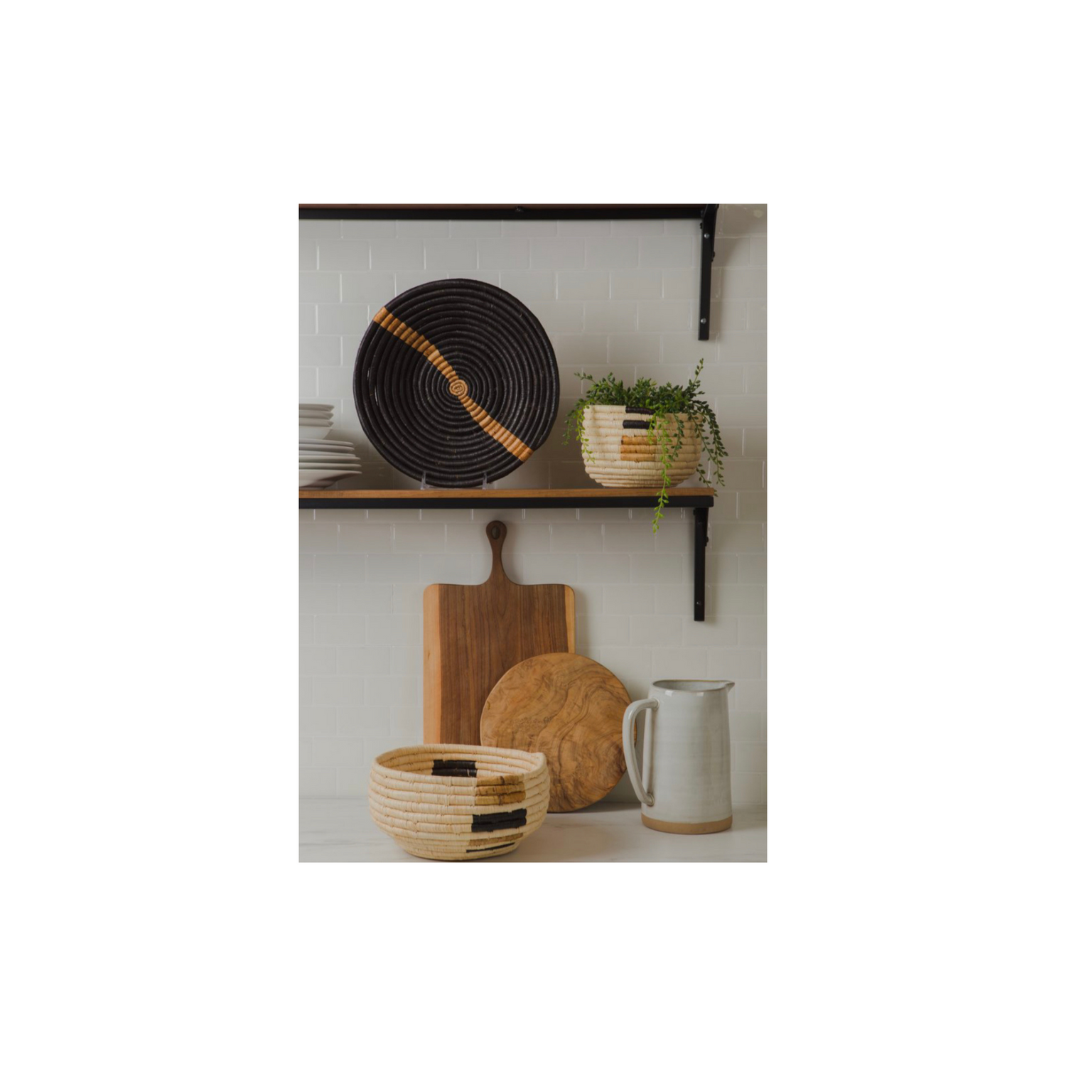 Shelves with woven baskets, bread boards against a white tile wall, ceramic pitcher and plant inside kazi woven basket